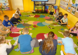 Grupa dzieci siedzi na dywanie, każde z dzieci układa historyjkę obrazkową.